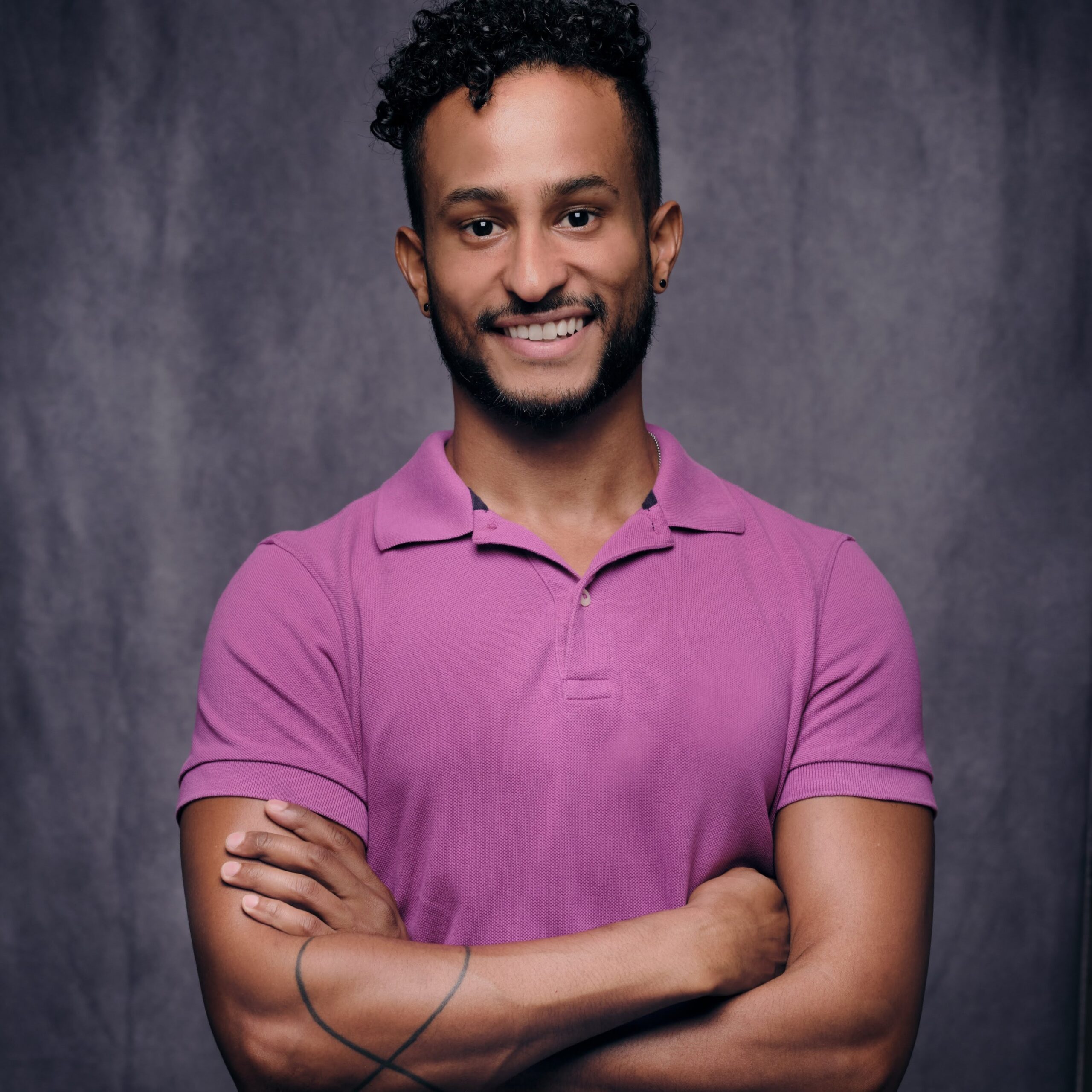 James--a Black and Latino man with brown skin, black curly hair and short beard, and dark brown eyes--is smiling at the camera in front of a blue-gray background. His smile is wide and crooked, he is wearing a magenta polo shirt, and his crossed arms show a tattoo on his right forearm.