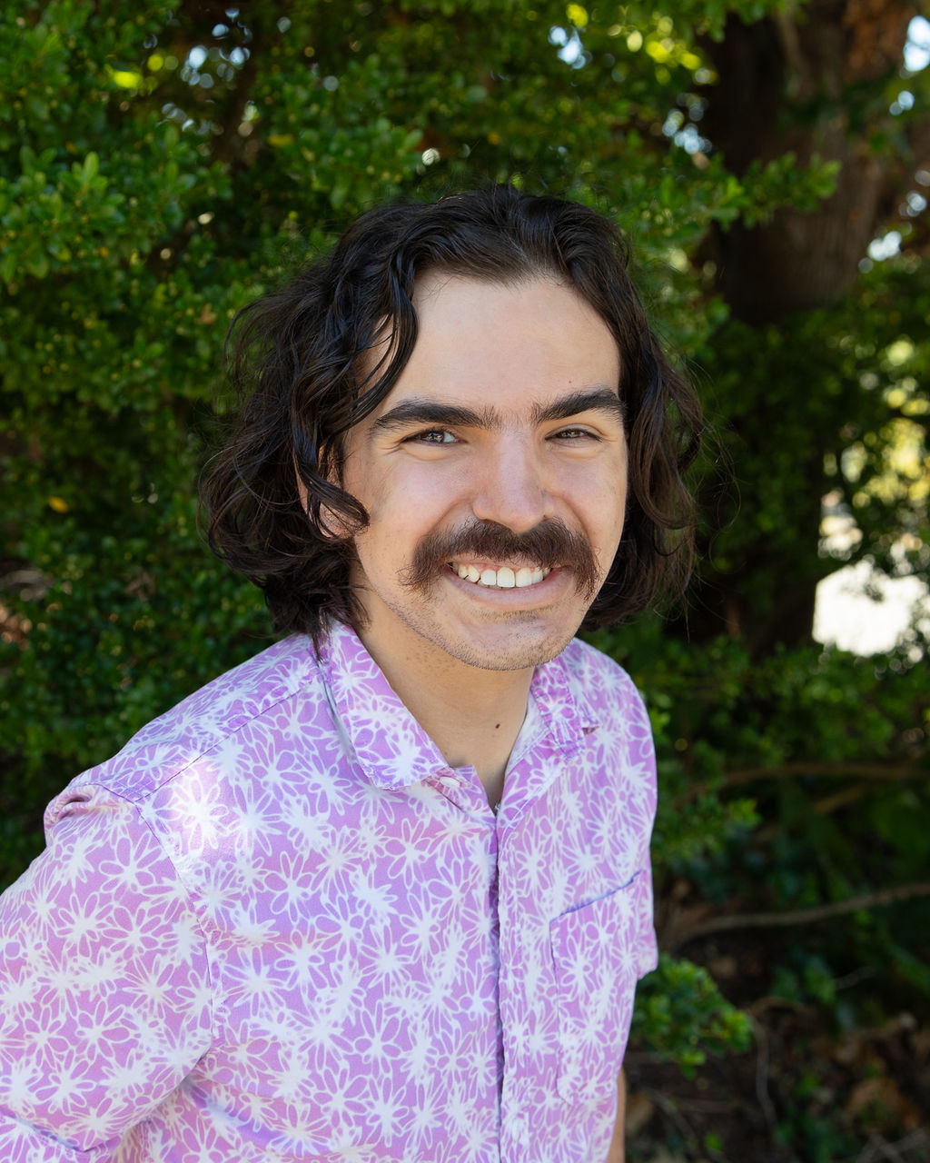 Image desc: Person with a medium length brown hair and a moustache wearing a pink floral short-sleeve button-up shirt. Person is smiling and standing in front of natural greenery