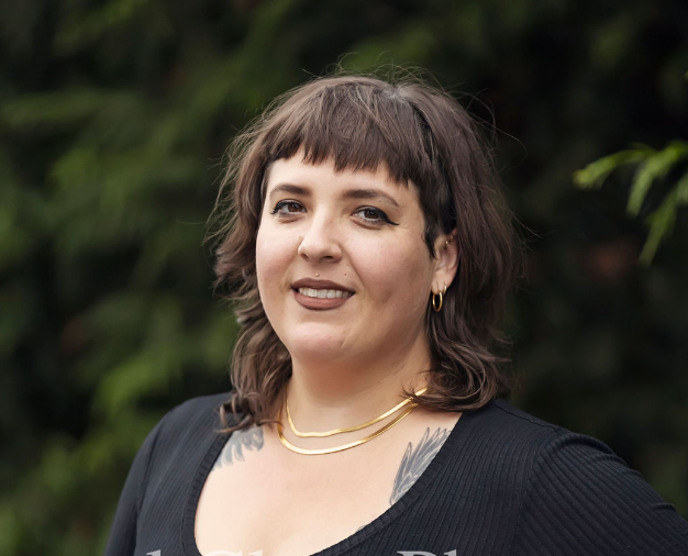 smiling smallfat brunette femme with tattoos wearing black shirt and gold necklace, brown lipstick in front of dark green foliage background