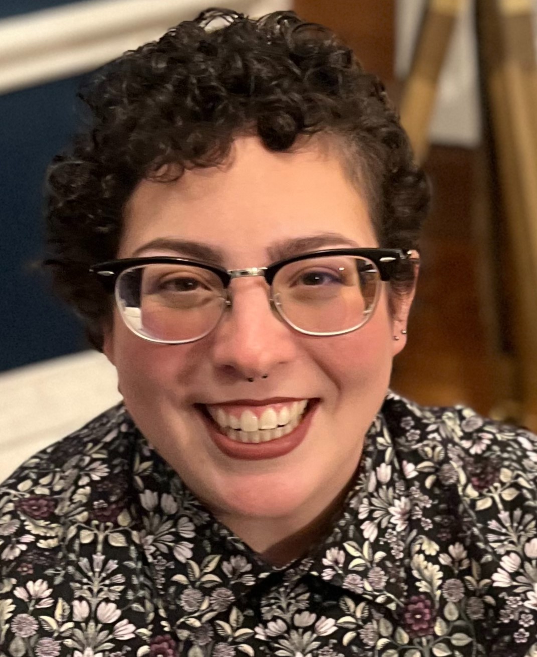 A white passing non-binary person with maroon lipstick, dark curly short hair, and black rimmed glasses smiles at the camera. They wear a button down shirt with white, maroon and black floral design.