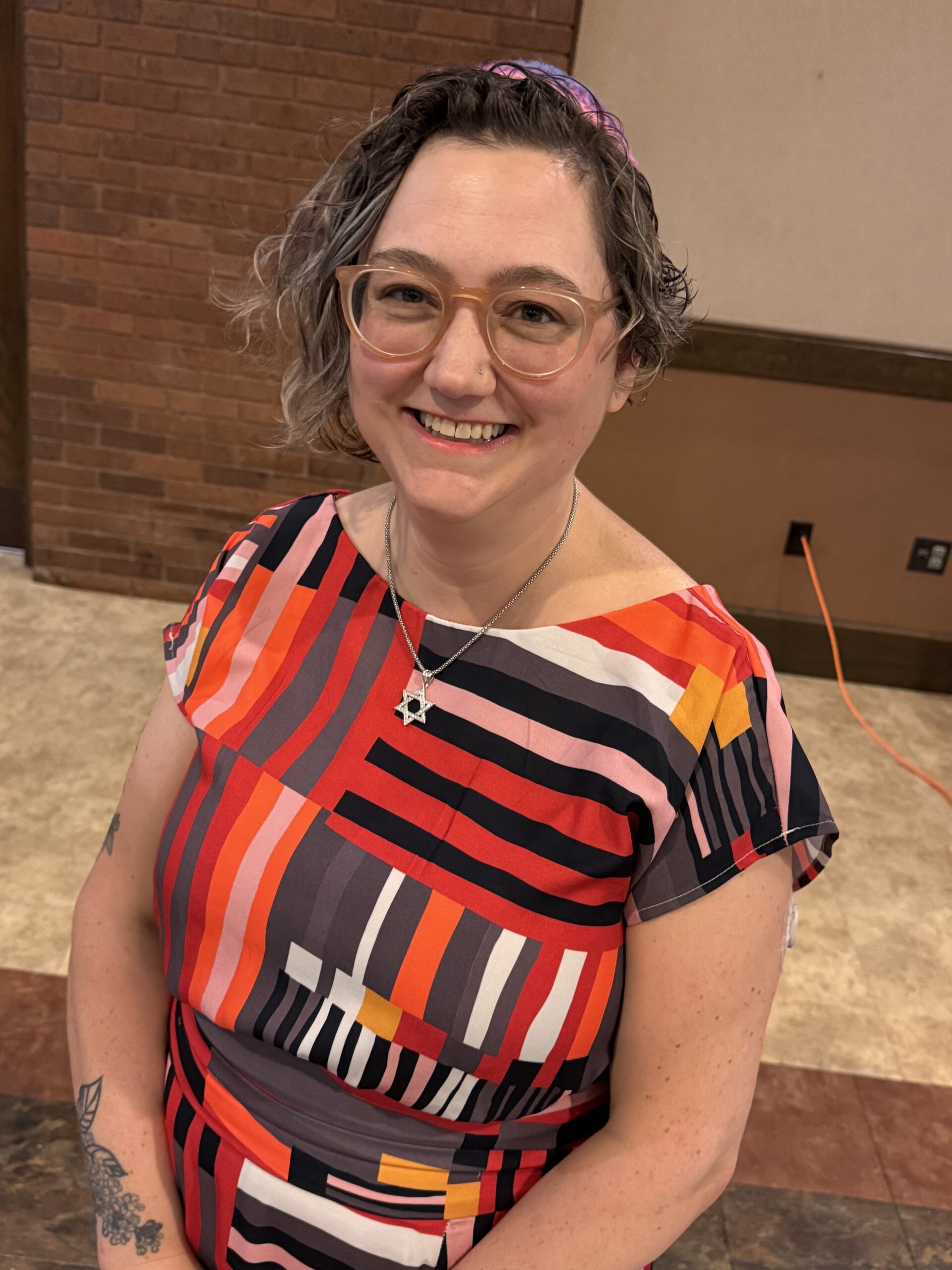 Rabbi Nikki, a white woman wearing a colorful dress and a rainbow crocheted kippa, smiles Rabbi Nikki, a white woman wearing a colorful dress and a rainbow crocheted kippa, smiles