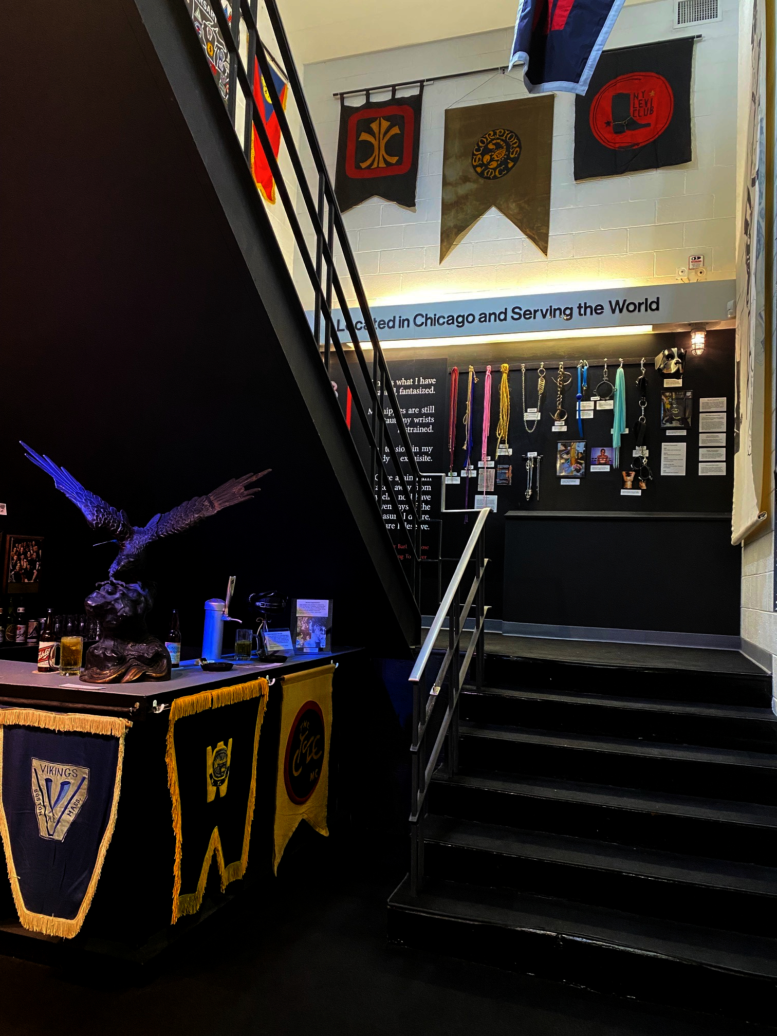 Hallway staircase with leather club banners, on display, an eagle statue, and a wall with rope, chains, fabric, and other bondage materials hanging from hooks.