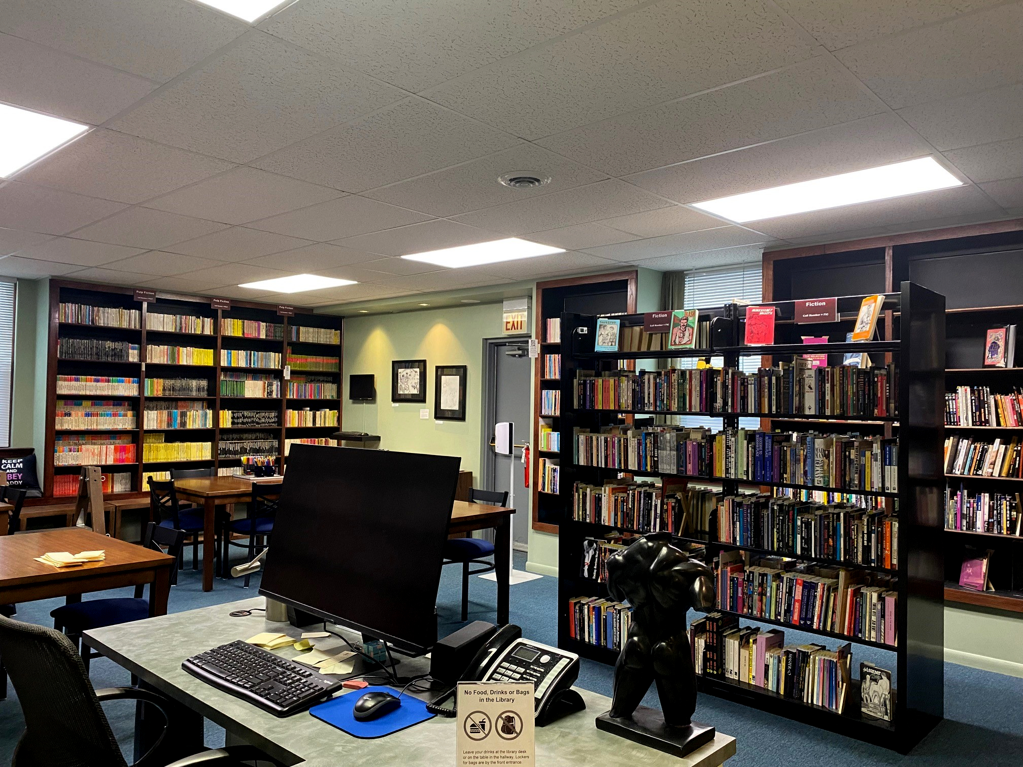 Library desk with computer, 1 free standing bookshelf visible behind with tables and bookshelves lining the walls.