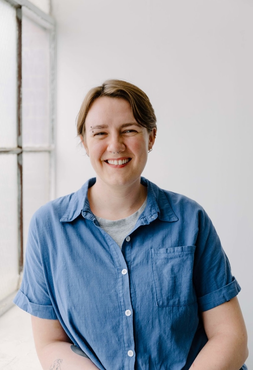 Courtney, a white non-binary person with short brown hair and brown eyes, is smiling with teeth showing and leaning against a window sill in a bright, white room. They are wearing a blue button-up short-sleeve shirt, silver septum, eyebrow and ear piercings, and they have tattoos on their forearm.