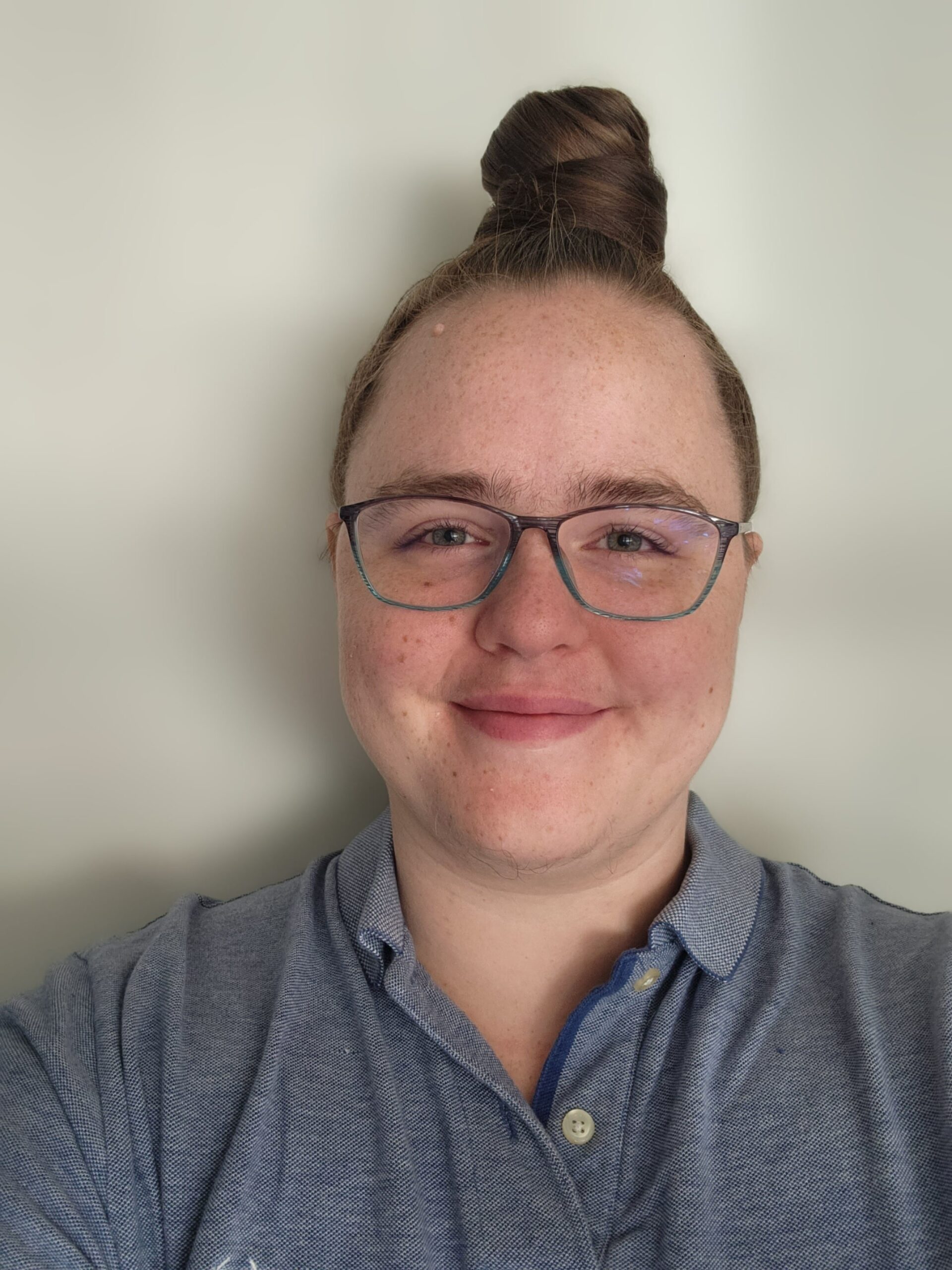 An image of a white, kinky, non-monogamous, neurodivergent, queer, transgender non-binary person with his hair in a bun, gently smiling.