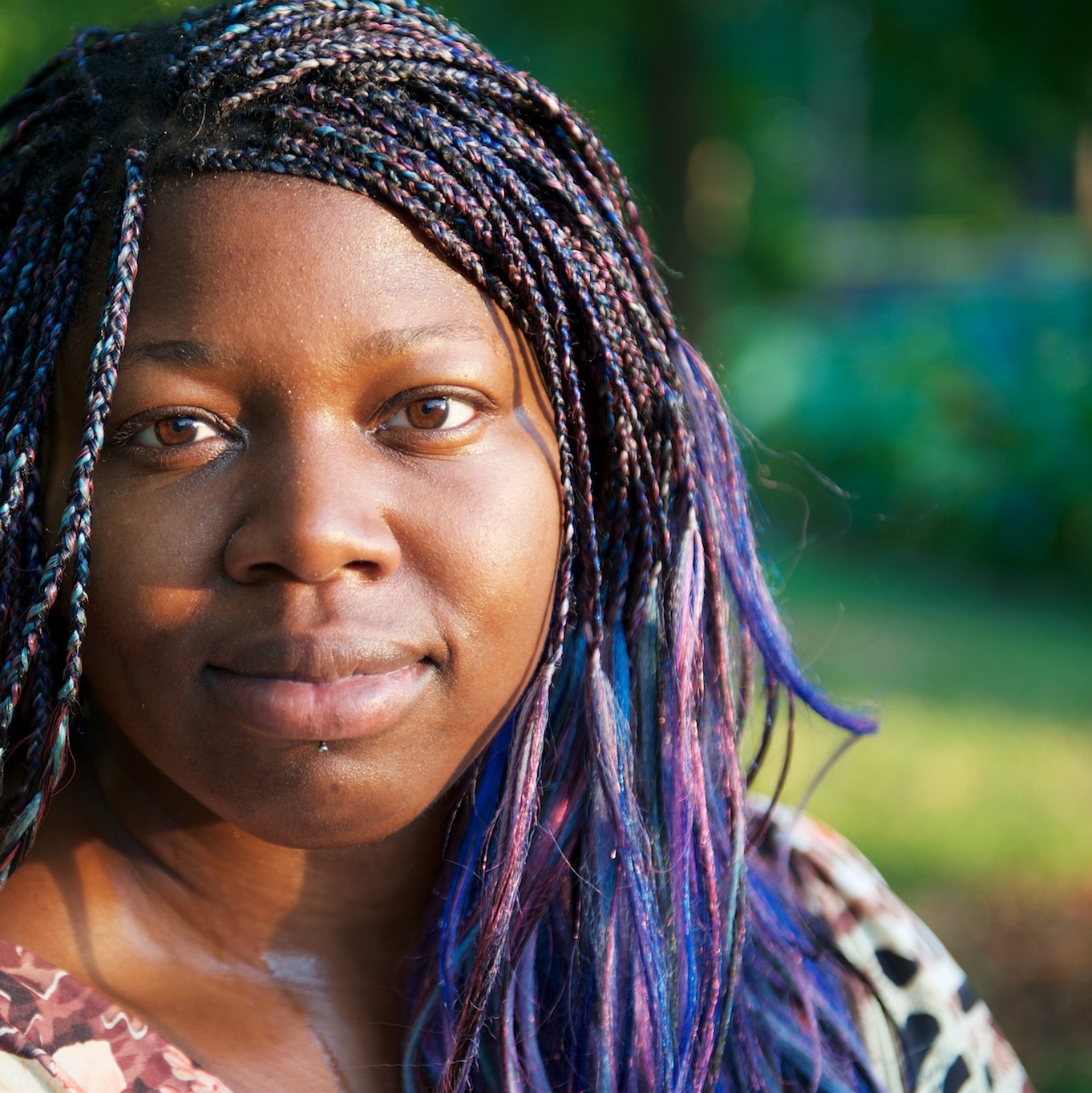 Brown-skinned human with purple braids looking at the camera with a slight smile.