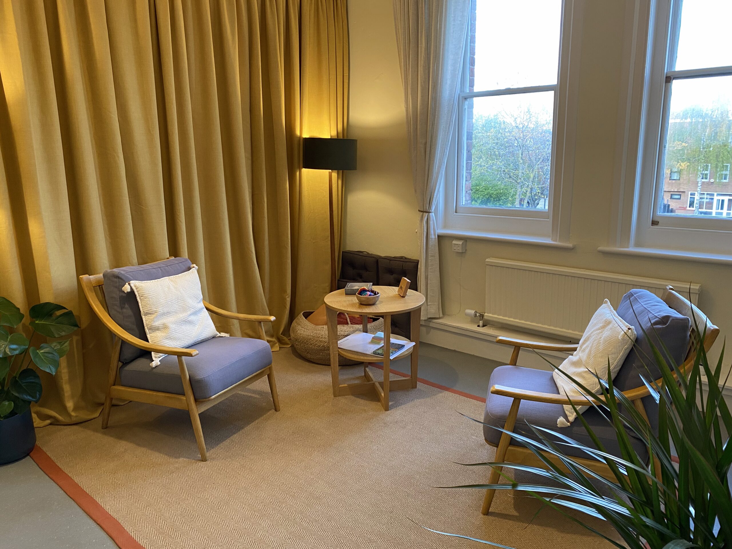 A comfortable practice room, with two armchairs, warm lighting, big windows and a natural carpet. There are green plants in the fore and background.