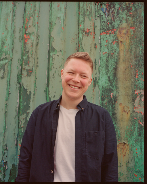 I am a white, trans non binary person with short hair, smiling, in a black shirt in front of a green multi-tone industrial background