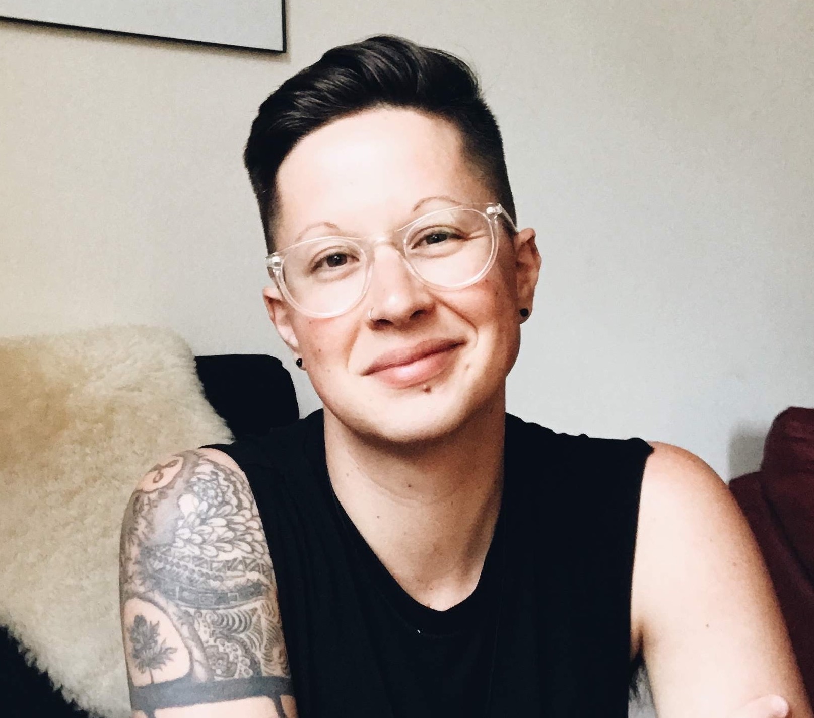 Korrine Wojcik is sitting in black chair with a fuzzy cream color blanket hanging over it. They have short dark hair, clear glasses, and are wearing a sleeveless black shirt. They have tattoos on their right arm. They are smiling at the camera. Korrine Wojcik is sitting in black chair with a fuzzy cream color blanket hanging over it. They have short dark hair, clear glasses, and are wearing a sleeveless black shirt. They have tattoos on their right arm. They are smiling at the camera.